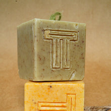 Load image into Gallery viewer, Two artisanal soap bars, one green with natural specks and one yellow, both featuring a distinctive embossed 'T' design.