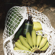 Load image into Gallery viewer, Coriander Seed Omega Body Oil bottle surrounded by bananas and a watermelon in a net bag.