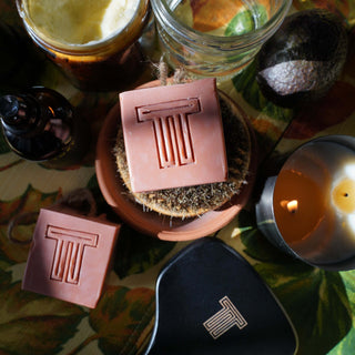 A bar of soap on a spoon, with a candle and a round tin container with a lid in the background, all placed on a colorful patterned surface.