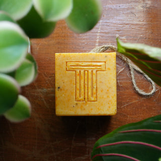 Dragon Fruit, Papaya & Carrot Soap cube surrounded by green leaves on a wooden surface.