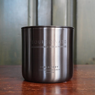 A silver stainless steel candle container with the label 'SOURSOP 430 SCENTED CANDLE' on a wooden surface with a metallic background.
