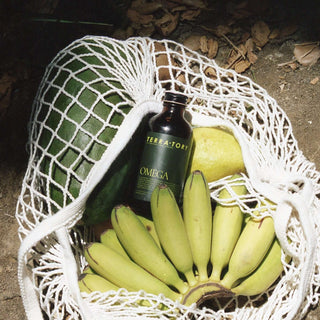 Coriander Seed Omega Body Oil bottle surrounded by bananas and a watermelon in a net bag.